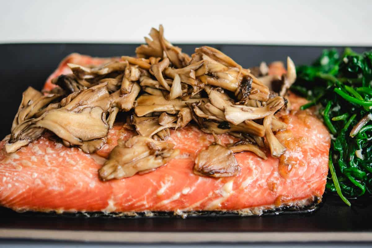 Sockey salmon filet with maitake mushrooms and wilted greens on a black rectangular plate.
